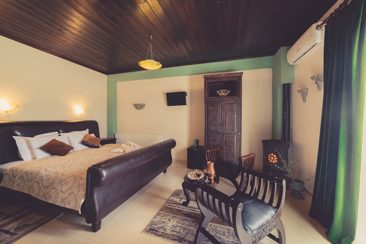 Quarto com cama, teto de madeira, radiador, aquecedor fixado na parede, pequena mesa com planta e uma cadeira.