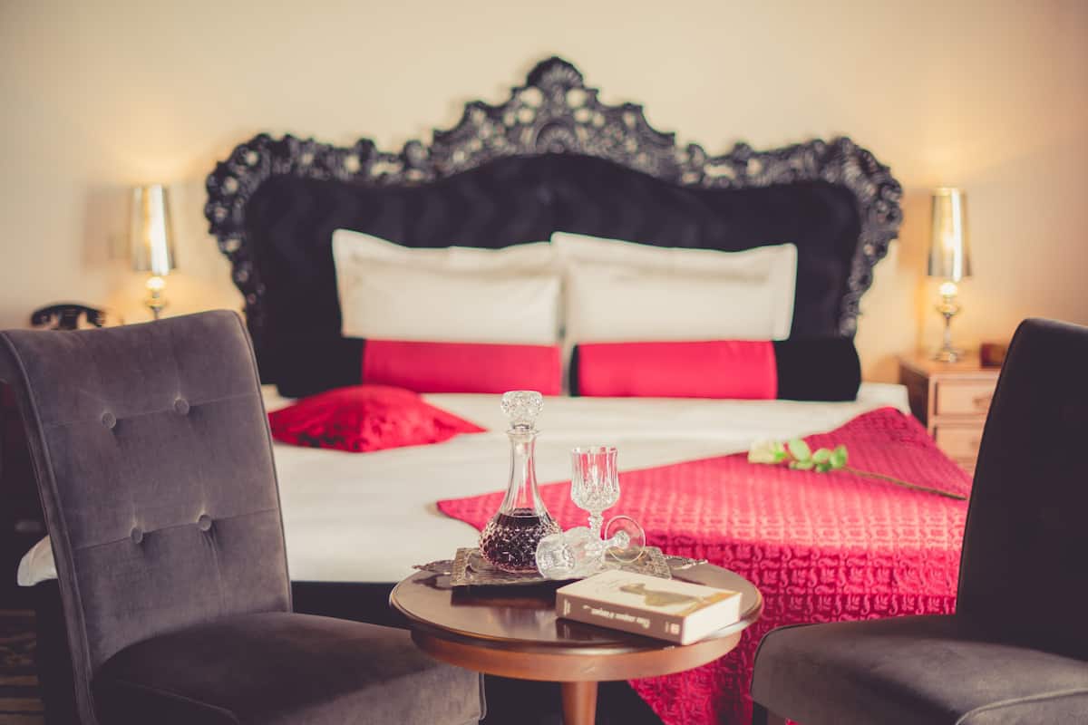 Uma cama bem arrumada com cabeceira preta, travesseiros vermelhos e brancos, e um cobertor vermelho. Há uma pequena mesa com um livro, um copo e uma garrafa. Duas cadeiras estão posicionadas em frente à cama.