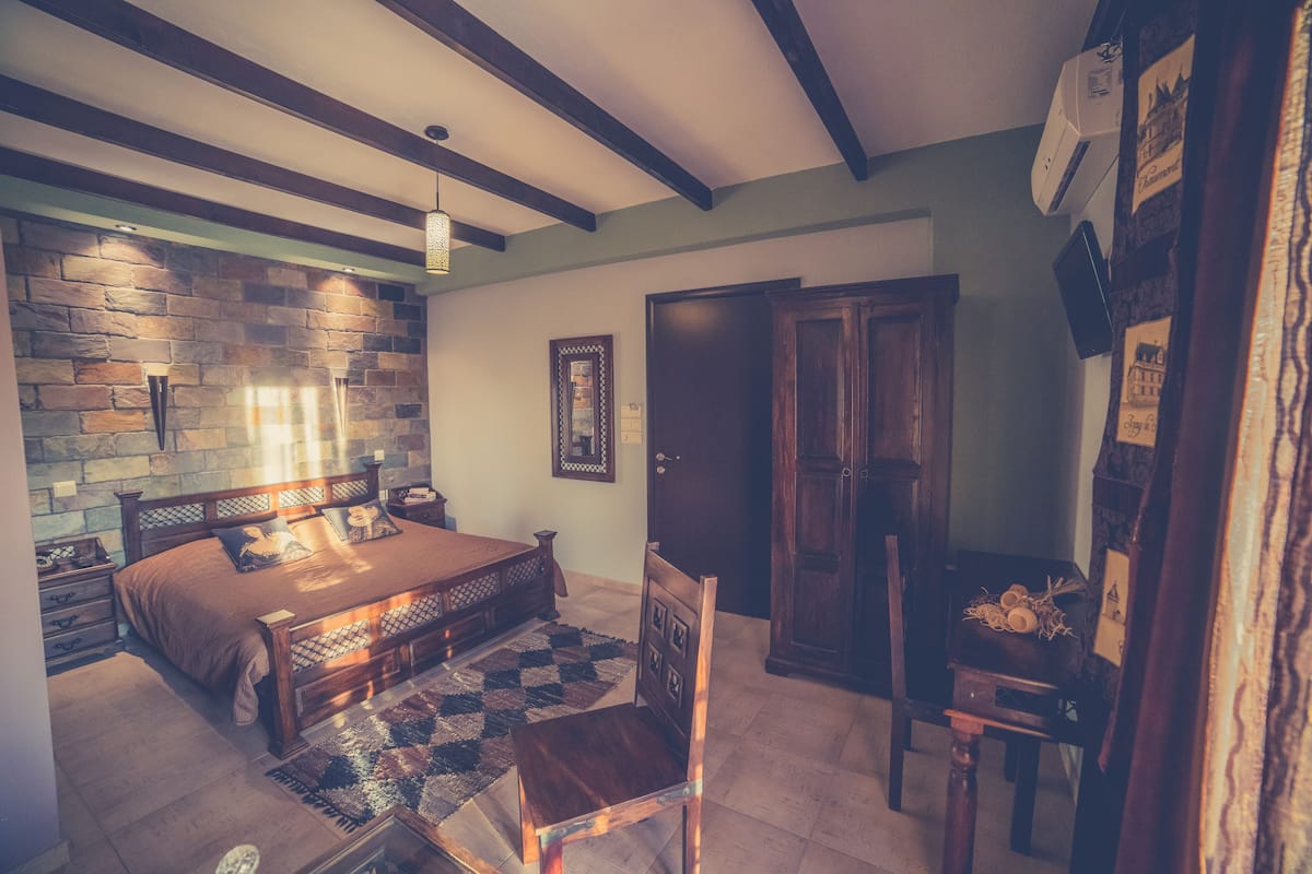 Quarto com cama, cabeceira de madeira, uma cadeira de madeira, uma mesinha, ar condicionado fixado na parede e uma porta.