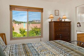 A bedroom with a large window, a bed with patterned bedding, a wooden dresser, and a lamp.