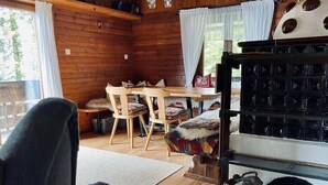 Ein gemütliches Blockhaus-Interieur mit Holzwänden, einem Essbereich mit Stühlen und Tisch, einem Kamin mit Stauraumschränken und Blick ins Freie durch große Fenster.