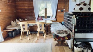 Ein gemütliches Blockhaus-Interieur mit Holzwänden, einem Esstisch aus Holz, zwei Stühlen, einer Bank, einer Chandelüster, einem Fenster mit Vorhängen und einem Regal mit Aufbewahrungsboxen.