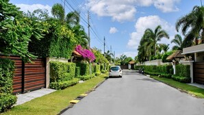 A residential street with a white car parked on the side, surrounded by lush greenery and palm trees. - Busaba Villa 7 - 3 Bedroom - Pool (Tambon Nong Kae)
