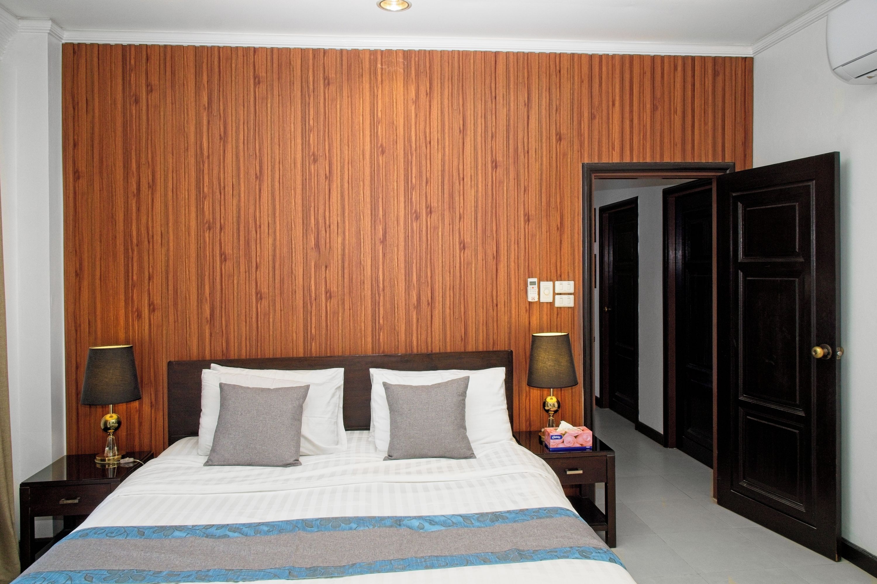 Ein Hotelzimmer mit einer Wandpaneele aus Holz, einem Bett mit weißer Bettwäsche und zwei Kissen, zwei Nachttischlampen, einem Nachttisch und einer Tür zu einem anderen Raum.