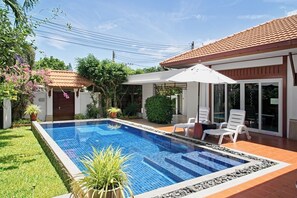 Une piscine dotée d’une terrasse carrelée, entourée d’un jardin où poussent différentes espèces de plantes, et un espace patio aménagé avec des chaises et un parasol.