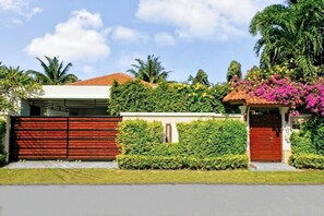 A residential property with a red garage door, a white building with a tiled roof, and a well-maintained garden with various plants and flowers. - Busaba Villa 7 - 3 Bedroom - Pool (Tambon Nong Kae)