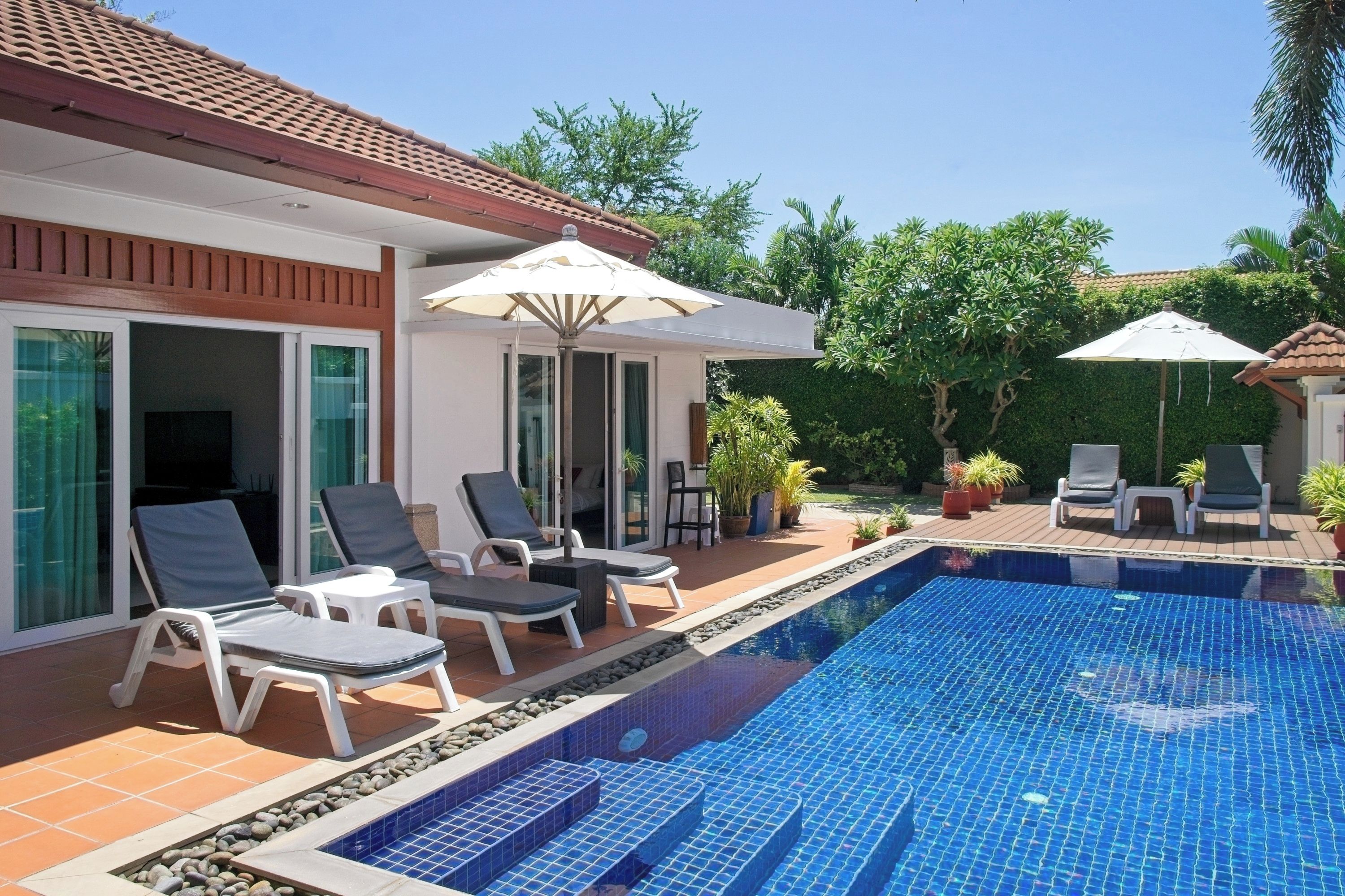 A swimming pool with lounge chairs and umbrellas, surrounded by a garden and a building with large windows.
