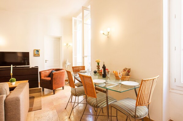 A dining area with a glass table, wicker chairs, and a television in the background. - LA CITTA VECCHIA AP2021 by RIVIERA HOLIDAY HOMES (Villefranche-sur-Mer)