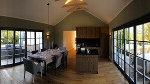 Een ruime eetruimte met een lange tafel gedekt voor zes personen, grenzend aan een keuken met houten kasten en een groot raam.