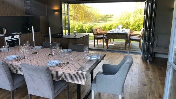 Een eethoek met een tafel voor zes personen, een keuken met moderne apparatuur, en uitzicht op een tuin door grote schuifdeuren.