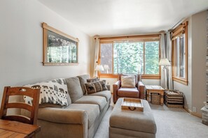Un salon chaleureux avec un canapé, un fauteuil et un pouf, une table d’appoint en bois, une lampe et une fenêtre donnant sur des arbres.