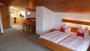 A bedroom with a wooden bed, a small dining table, and a kitchen area in the background.