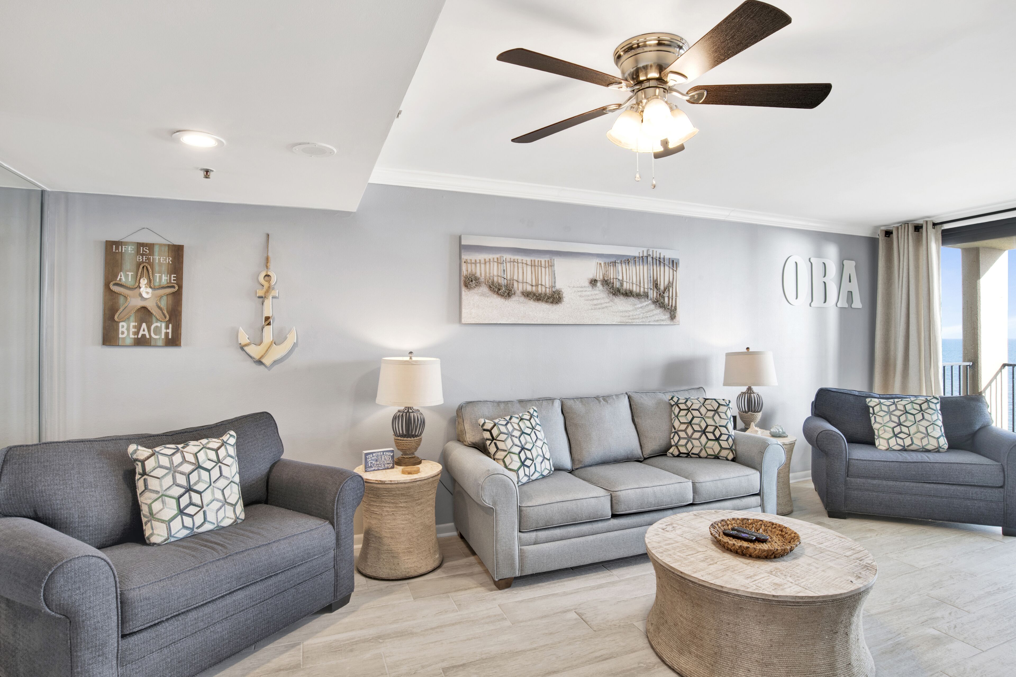 A modern living room with grey sofas, patterned cushions, a ceiling fan, and a large window with curtains.