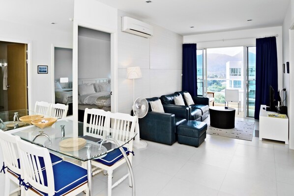 A modern hotel room with a glass dining table, white chairs, a blue sofa, and a balcony with a view of buildings. - SEACRAZE D709 - 2 Bedroom - Near Beach (Tambon Nong Kae)