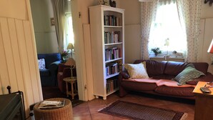 Una sala de estar acogedora con un sofá de cuero marrón, una estantería blanca, una mesita de mimbre y suelo de baldosas.