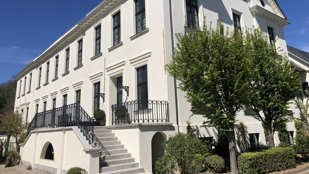 Un edificio grande y blanco con fachada simétrica, numerosas ventanas y una escalera central que lleva a la entrada.