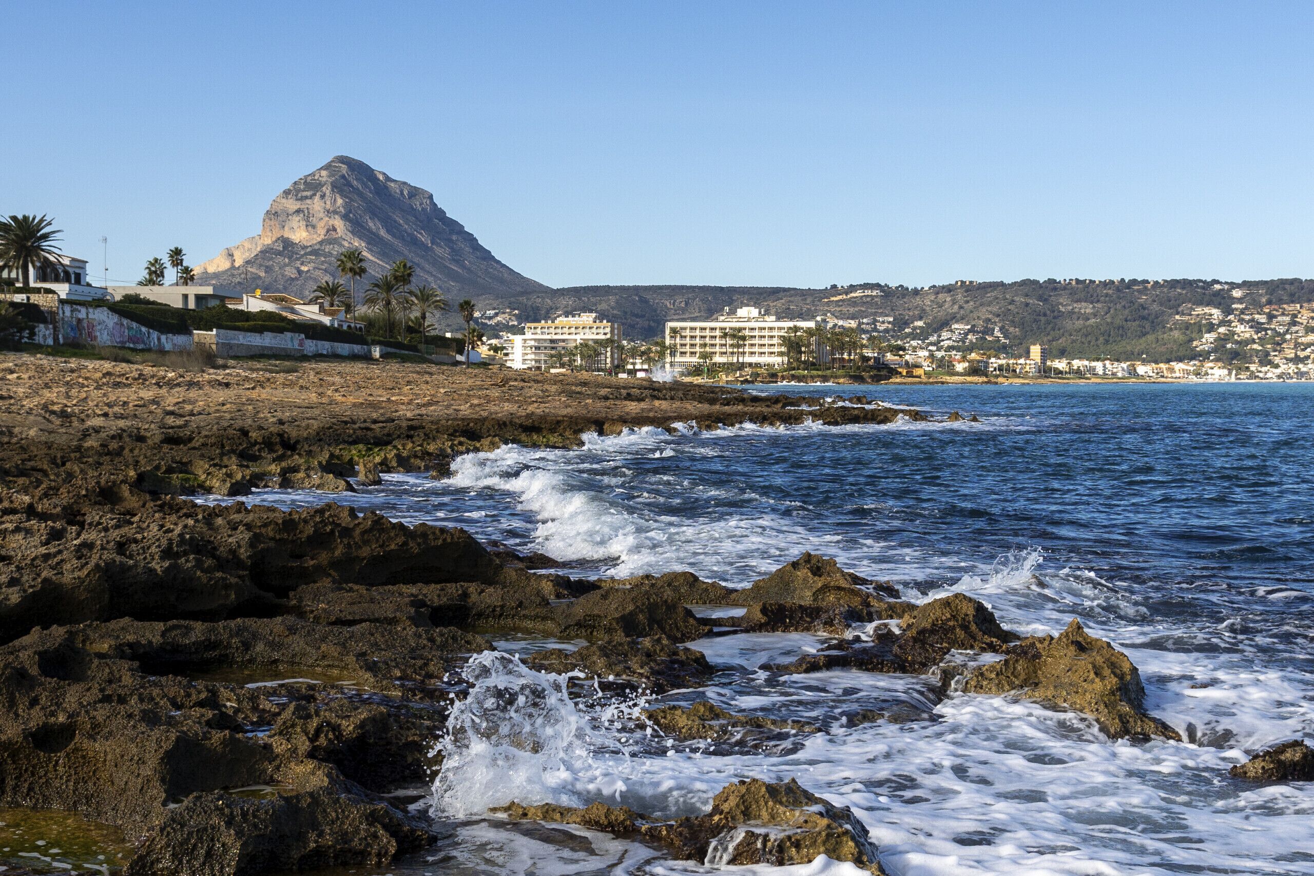 A coastal hotel with rocky shoreline, palm trees, and a mountain in the background.