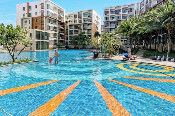 A swimming pool with a tiled pattern, surrounded by multi-story buildings and palm trees.