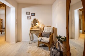 A cozy room with a wicker chair, a small desk with a lamp, and a mirror. There are framed pictures on the wall and a fluffy rug on the floor.