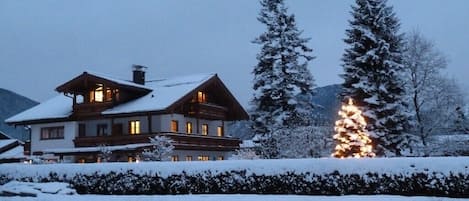 A large, multi-story chalet with a snow-covered roof and a lit Christmas tree in the background.