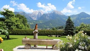 Ein malerischer Garten mit einer Holzbank und einer Statue, umgeben von üppiger Vegetation und Bergen im Hintergrund.