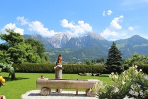 A scenic garden with a wooden bench and a statue, surrounded by lush greenery and mountains in the background. - Ferienwohnung Hochkalter, 2 bis 4 Personen, 82 qm (Schönau am Königssee)