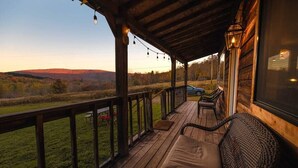 Una terraza de madera con un banco acolchado y una silla de mimbre, con vistas a un campo cubierto de césped y colinas lejanas.