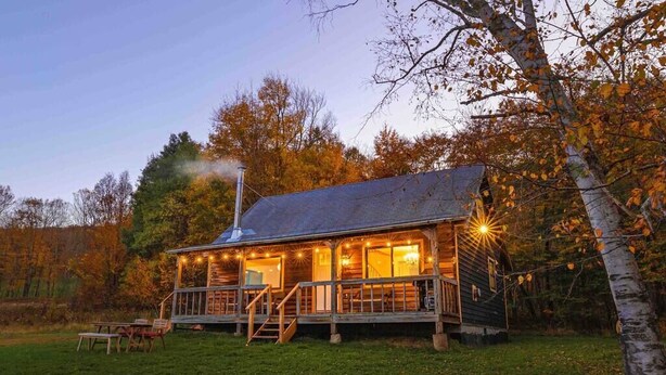 Una cabaña de madera con un porche, una chimenea y una pequeña terraza con mobiliario exterior.