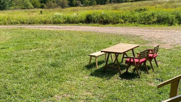 Un área de estar al aire libre con una mesa y sillas de madera en un campo cubierto de césped, con una montaña y bosque al fondo.