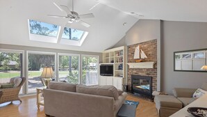 Un salón espacioso con un sofá grande, chimenea, televisor y estantería. Cuenta con ventanas amplias y un ventilador de techo.