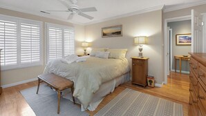 Un dormitorio con cama, lámparas de noche, un banco, ventilador de techo y vista al pasillo.