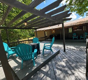 A wooden deck with turquoise chairs and a table, a covered area with a pergola, and a building with a metal roof in the background. - "Cottage @ JP Coleman"-perfect getaway at Pickwick (Iuka)