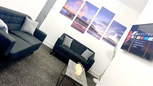 Una sala moderna con un sofá gris, un sillón más pequeño y una mesa de centro. Hay tres cuadros en la pared que muestran una noria y una puesta de sol.
