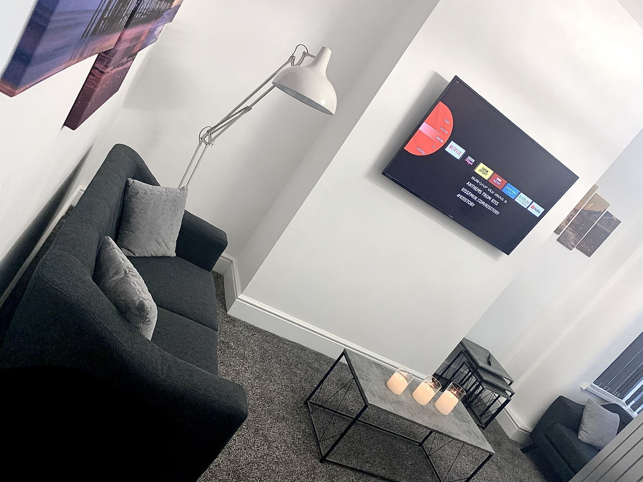 Een moderne woonkamer met een donkergrijze bank, een vloerlamp, een salontafel en een flatscreen-tv aan de muur, waarop verschillende streamingdiensten worden getoond.