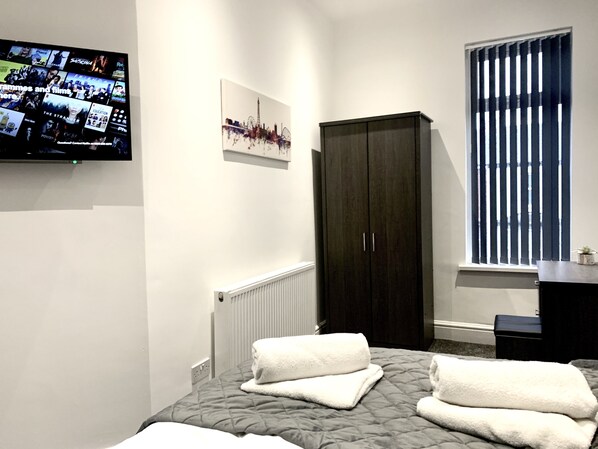 A bedroom with a bed, a dark wardrobe, a radiator, a window with blinds, and a TV displaying a program.