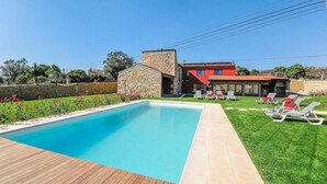 Una piscina con muelle de madera, un edificio de piedra y una casa con techo rojo, césped y sillones de descanso.