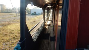 L’intérieur d’un wagon de train, avec un banc en métal le long du couloir, une rampe d’appui en métal et une vue sur un espace extérieur comprenant un bâtiment et quelques voitures garées.