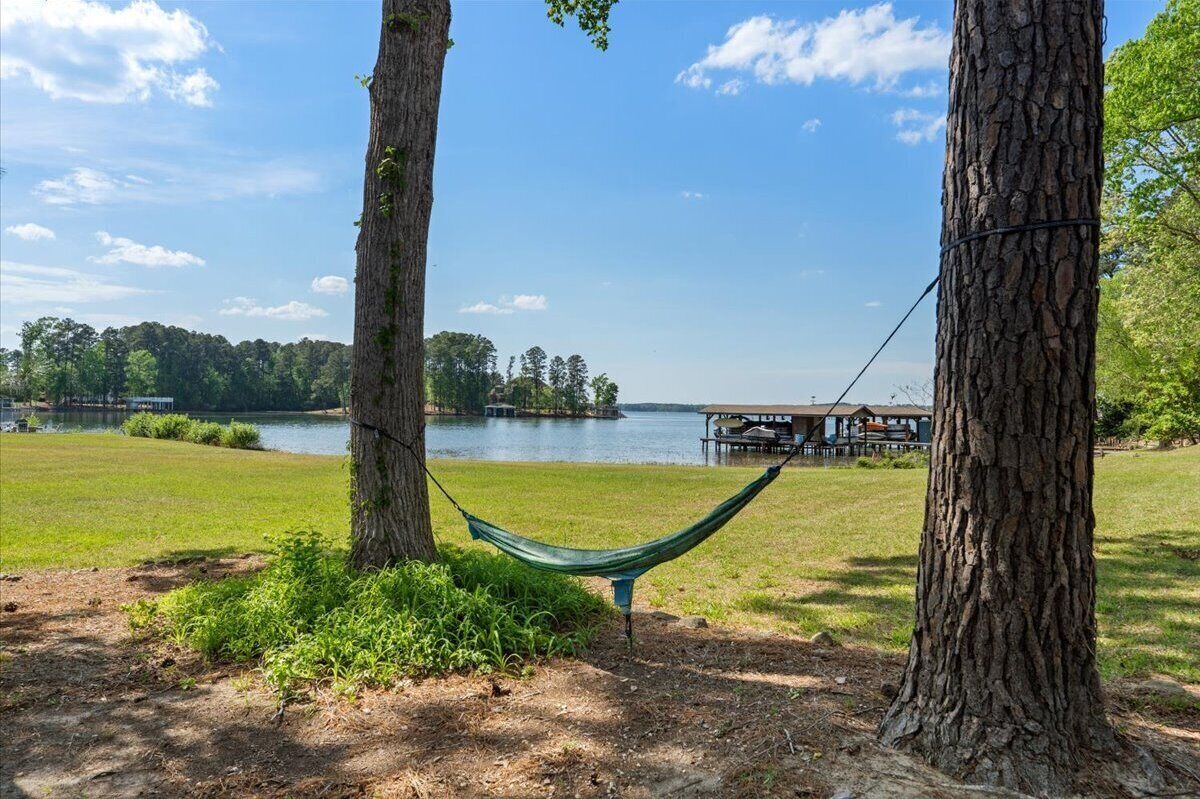 A hammock between trees by a lake with a dock and buildings in the background.