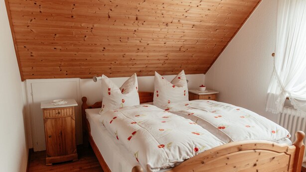Una cama de madera con techo inclinado, ropa de cama blanca con diseños florales, una mesita de noche y una lámpara de noche.