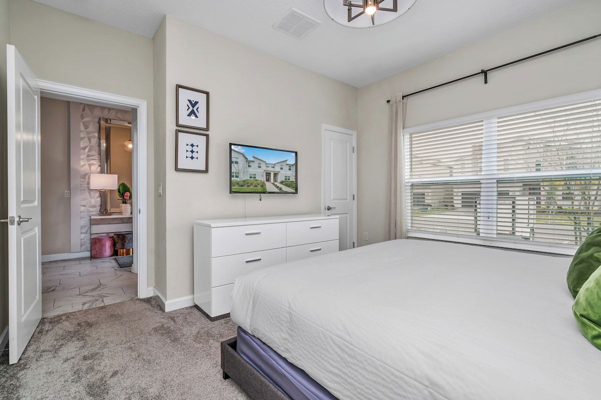 A bedroom with a bed, a dresser, a television, and a window with blinds.