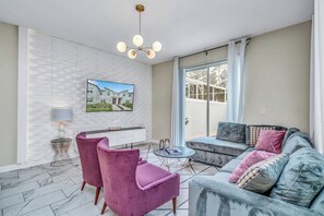 Un salon moderne avec un mur en briques blanches, un téléviseur à écran plat, un canapé gris avec des coussins, une table basse ronde et deux fauteuils roses.