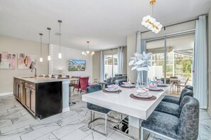 Une cuisine moderne dotée d’un îlot central, d’un coin repas avec une longue table et des chaises, et de grandes fenêtres laissant entrer la lumière naturelle.