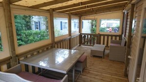 Un espace extérieur abrité, doté d’une terrasse en bois, d’une table avec des bancs et offrant une vue sur une maison à travers les fenêtres.
