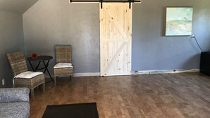Una habitación con una puerta corredera de madera, dos sillas de mimbre, una mesita con una flor, un sofá gris y un cuadro en la pared.