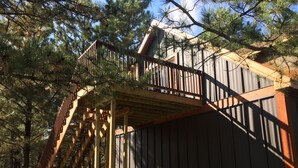 Una escalera de madera que lleva a una terraza con baranda de metal, rodeada de árboles.