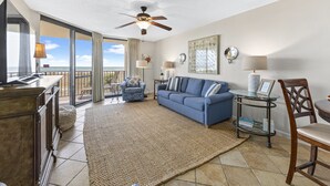 A spacious living room with a large blue sofa, a wooden coffee table, and a ceiling fan. There is a large window offering a view of the ocean. - Phoenix VII 215 | Beachfront Condo | Resort Amenities | My Beach Getaways (Orange Beach)