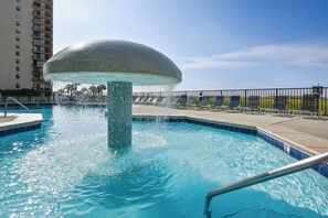 A swimming pool with a water fountain feature, surrounded by lounge chairs and a fence.