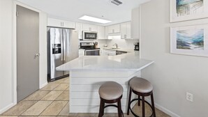 A modern kitchen with white cabinetry, a central island with stools, a refrigerator, microwave, and stove. - Phoenix VII 215 | Beachfront Condo | Resort Amenities | My Beach Getaways (Orange Beach)