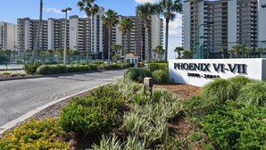 A sign for Phoenix VI-VII apartments with a view of tall residential buildings and palm trees. - Phoenix VII 215 | Beachfront Condo | Resort Amenities | My Beach Getaways (Orange Beach)