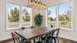 A dining area with a wooden table and chairs, large windows, and a hanging light fixture. - Juniper Ranch - Golf Cart Included | Sleeps 10 (Powell Butte)